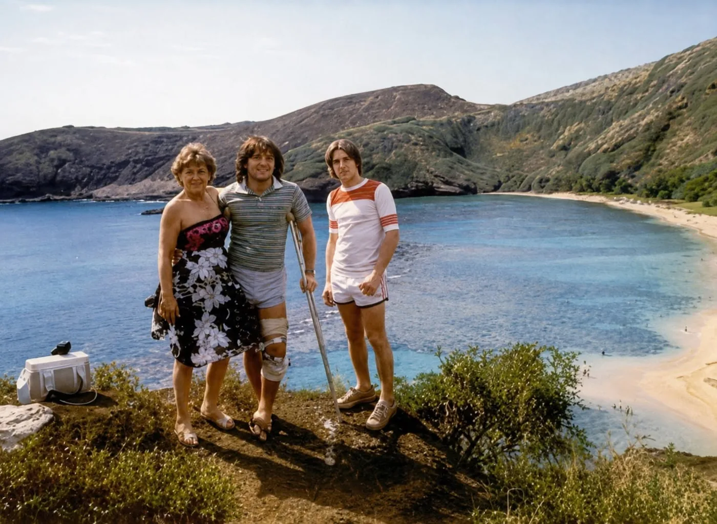 Three people overlooking a Hawaiian bay, with the center person using crutches and wearing a knee brace